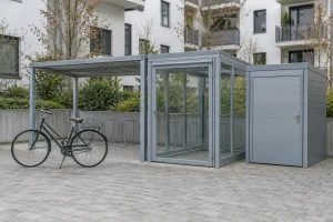 Abri vélo collectif gris avec structure en métal et porte verticale, entouré d’un vélo et d’un toit et de façades vitrées.