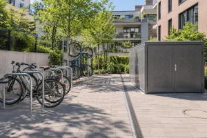 Espace extérieur avec plusieurs vélos fixés sur des porte-vélos et un cabanon en métal sur une allée pavée.
